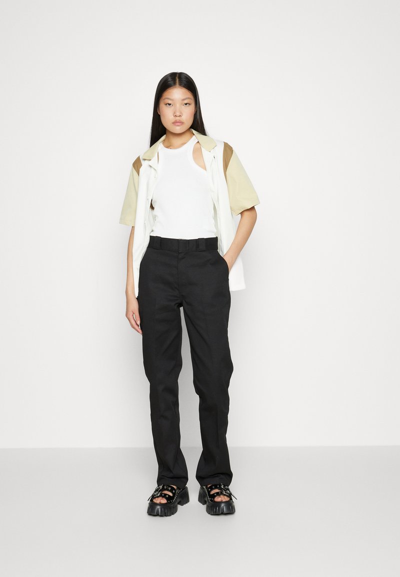 Débardeur blanc avec découpes, superposé avec une chemise à manches courtes vert clair et marron, associé à un pantalon noir taille haute et des sandales épaisses.