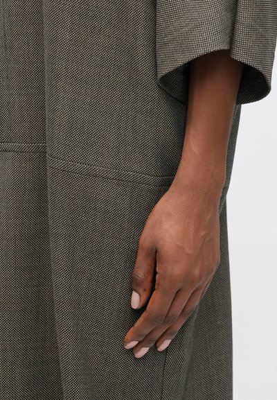 Szürke textúrájú anyagból készült, laza szabású ruha, széles ujjal és varrási részletekkel, mellette egy kéz látható, amelynek körmei semleges árnyalatúak.