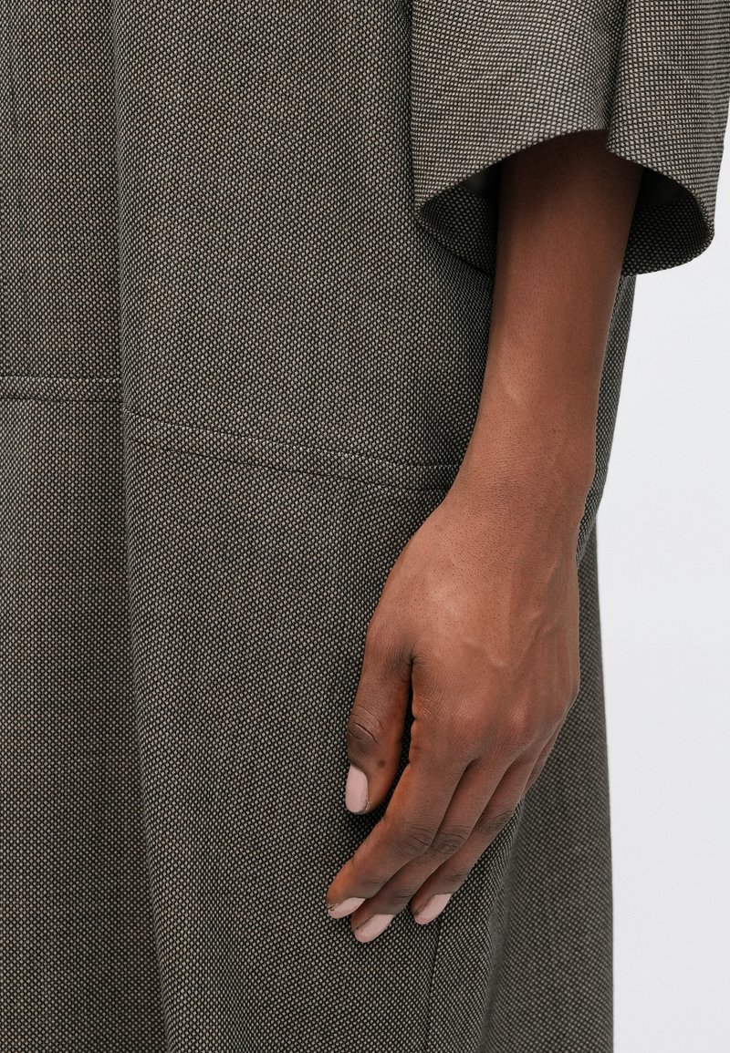 Vêtement en tissu texturé gris avec une coupe décontractée, présentant de larges manches et un détail de couture, à côté d'une main avec des ongles de teinte neutre.