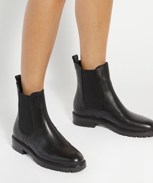 Jambes portant des bottines Chelsea en cuir noir avec panneaux latéraux élastiques et languettes de tirage, debout sur un sol blanc.