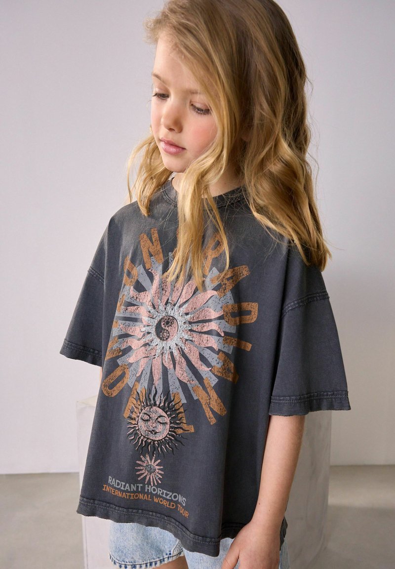 Junges Mädchen mit langen blonden Haaren, das nach unten schaut, trägt ein dunkles, übergroßes T-Shirt mit Sonnen- und Textgrafik, kombiniert mit hellen Jeansshorts.