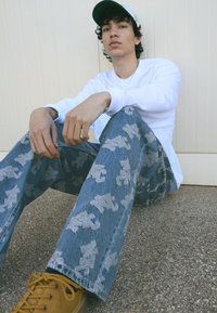 Světlé modré džínové kalhoty s texturovaným vzorem, zdobené šedým abstraktním designem. Vytvořené v kombinaci s bílou košilí s dlouhým rukávem a žlutými botami.