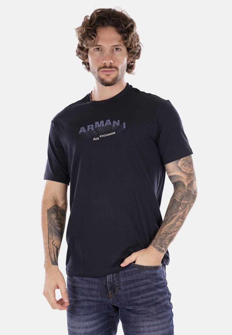 T-shirt nera a maniche corte in cotone con stampa grafica "ARMANI EXCHANGE" in blu e grigio. Modello mostrato con tatuaggi e jeans blu.