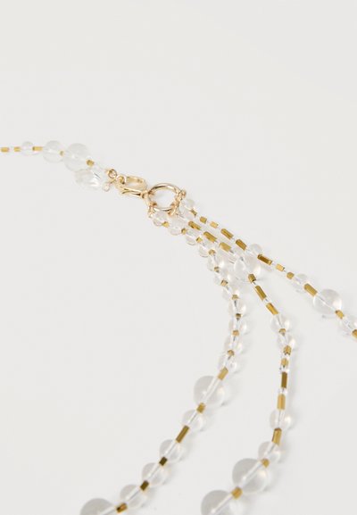 Κούμπωμα κολιέ με χρυσό αστακό και κρίκο, προσαρτημένο σε σειρές από διάφανα στρογγυλά χάντρες και μικρές χρυσές κυλινδρικές χάντρες σε λευκό φόντο.