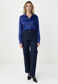 Satenowa granatowa koszula z długim rękawem w połączeniu z ciemnoniebieskimi spodniami o wysokim stanie, które mają przednie kieszenie oraz kontrastowe pomarańczowe przeszycia.