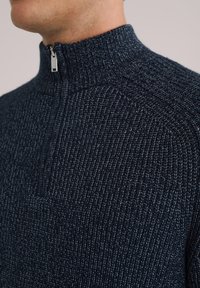 Jūras spēku ceturtdaļas rāvējsvērs ar ribotu tekstūru un smalku tonālo akcentu. Iezīmē augstu apkakli un metāla rāvējsvēršanas vilkmi.