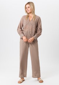 Blouse beige brodée à manches longues et détails découpés, associée à un pantalon beige ample avec des ourlets décoratifs et un tissu léger.