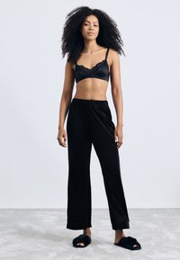 Musta satiininen bralette, jossa on pitsiyksityiskohtia, yhdistettynä löysiin mustiin pehmeisiin loungereisiin ja pehmeisiin mustiin tossuihin. Yksinkertainen, rento istuvuus kauttaaltaan.