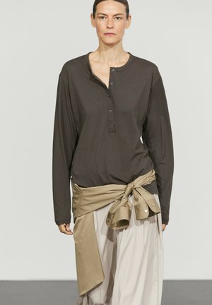 Woman wearing dark long-sleeve henley shirt with beige jacket tied around waist and light-colored loose pants, standing against plain background.
