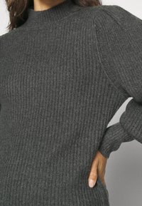 Frau, die einen dunkelgrauen, gerippten Strickpullover mit Stehkragen trägt, mit einer Hand an der Hüfte vor einem neutralen Hintergrund.