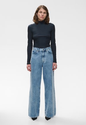 Femme aux cheveux mi-longs portant un pull noir à col roulé à manches longues et un jean bleu large taille haute, debout devant un fond uni.