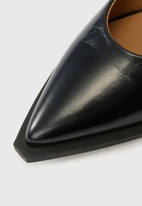 Μαύρο δερμάτινο παπούτσι με μυτερή μύτη και ελαφρώς τετράγωνη γόμα στη σόλα, σε ανοιχτό φόντο.