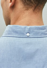 Jasnoniebieski kołnierzyk koszuli z krótkim rękawem i zapięciem na guzik. Tkanina wydaje się gładka i lekko teksturowana. Szwy widoczne w pobliżu krawędzi kołnierza.