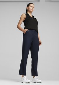 Top sin mangas negro con cuello, combinado con pantalones azul marino. Los pantalones tienen un diseño aerodinámico y bolsillos laterales. Zapatillas deportivas blancas.