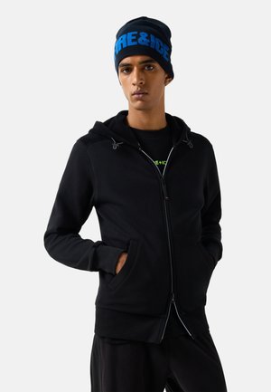 Ung mand iført en sort lynlåshættejakke og en sort hue med blå "FIRE&ICE" tekst, stående med hænderne i lommerne mod en ensfarvet baggrund.