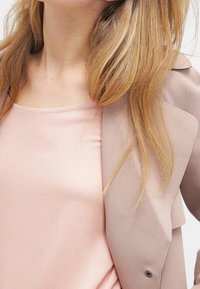 Blazer beige sopra una maglietta rosa chiaro. Il blazer presenta una texture morbida e una vestibilità su misura con chiusura a un bottone. Capelli acconciati in modo casual.
