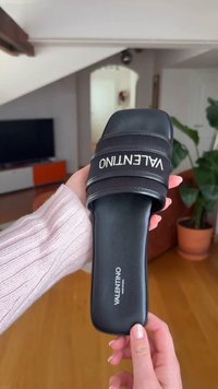 Χέρι κρατά ένα μαύρο σανδάλι Valentino slide με λευκό λογότυπο, εσωτερικός χώρος με ξύλινο πάτωμα, φυτά και τηλεόραση στο φόντο.