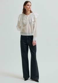 Kobieta ubrana w przezroczystą białą bluzkę z falbankami, ciemne dżinsy z szerokimi nogawkami i czarne buty na obcasie w szpic, stojąca na jednolitym tle.