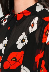 Zwarte bloes met bloemenprint met rode en witte klaprozen, een kraag en knoopdetails. Doorzichtige textuur met zachte draping.