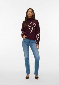 Burgundski pleteni pulover s turtleneck ovratnikom in kremnimi spiralami, v kombinaciji z svetlo modrimi džinsami. Ima dolge rokave in sproščen kroj.