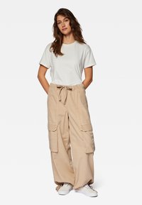 Beige Cargohose mit weitem Bein, seitlichen Taschen und verstellbarem Bund, kombiniert mit einem schlichten weißen T-Shirt mit kurzen Ärmeln und weißen Sneakern.