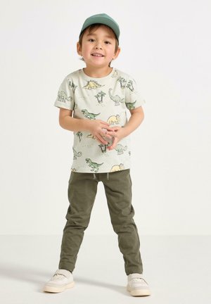 Nuori lapsi, jolla on dinosaurustehdellä painettu t-paita, oliivinvihreät housut, valkoiset lenkkitossut ja vihreä baseball-lippalakki, seisoo käsivarret ristissä.
