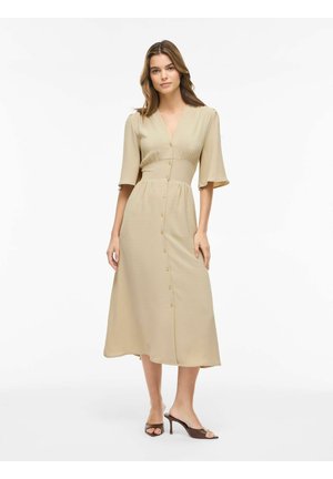 Kvinde iført en beige midikjole med knapper foran, trekvartlange ærmer og brune åbne hæle, stående mod en hvid baggrund.