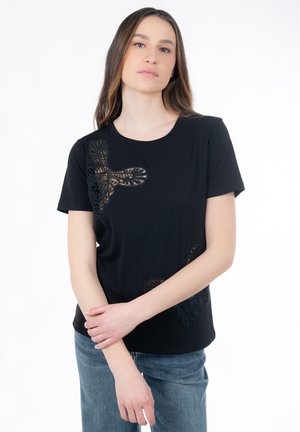 Jonge vrouw draagt een zwart topje met korte mouwen en kanten vlinderdetails, staat met armen losjes gekruist tegen een effen witte achtergrond.