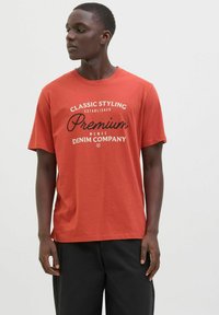 Raudonas medvilnės marškinėliai su trumpomis rankovėmis, su juodu spausdintu užrašu "Klasikinis stilius įkurtas Premium Denim Company." Paprastas apvalus iškirpimas.