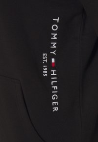 Sudadera negra con capucha que presenta texto bordado en blanco "TOMMY HILFIGER EST. 1985" con un detalle de logo en rojo y blanco. Tejido suave y texturizado.