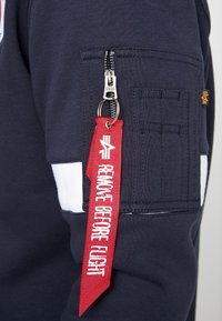 Sweat-shirt marine avec une poche zippée, des accents blancs et une étiquette rouge portant la mention "RETIRER AVANT LE VOL", attachée par un anneau en métal.