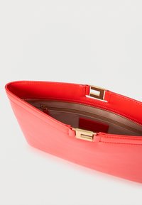 Φάκελος από δέρμα κοραλλί με ορθογώνιο χρυσό κούμπωμα, ανοιχτό πάνω μέρος που αποκαλύπτει το μπεζ εσωτερικό επένδυμα και εσωτερική τσέπη με φερμουάρ.