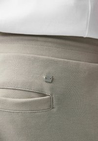 Pantalones de chándal gris claro con una tela texturizada, un bolsillo trasero y un pequeño detalle de logo cerca de la cinturilla.