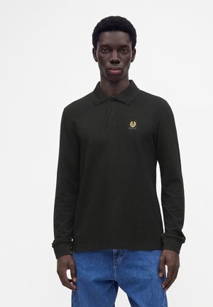 Mand iført en sort langærmet polo med en lille gul logo på brystet og blå jeans, stående mod en hvid baggrund.
