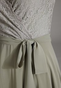 Corpetto in pizzo grigio chiaro con motivo floreale, abbinato a una gonna fluida verde. Presenta un fiocco legato in vita per la regolazione.