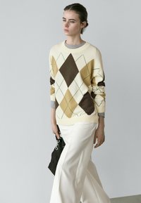 Junge Frau geht spazieren, trägt einen beigen Argyle-Pullover über einem grauen Hemd, weiße Hose und hält eine schwarze Handtasche vor einem schlichten Hintergrund.