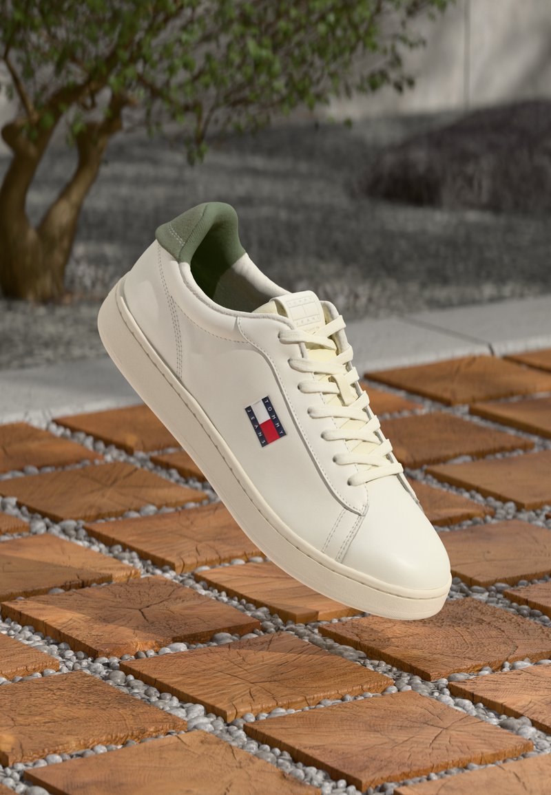 White leather sneaker with green heel tab and Tommy Jeans logo, floating above wooden square tiles with small gray pebbles.