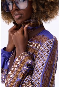 Kleurrijke blouse met een patroon in blauw en bruin, met ingewikkelde ontwerpen en een hoge kraag. De mouwen hebben een geplooid detail voor extra textuur.