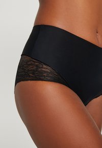 Mutandine nere con finitura opaca e liscia, decorate con pizzo lungo le gambe, per un contrasto e una texture interessanti. Design a vita media con retro senza cuciture.