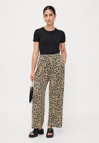 Zwarte top met korte mouwen en kleine studs, gecombineerd met losse beige broek met luipaardprint. Het model draagt zwarte platform sandalen en houdt een zwarte handtas vast.
