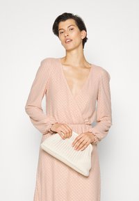 Vestido rosa, com padrão, com um profundo decote em V e mangas longas. Modelo segura uma carteira texturizada em tom off-white. Apresenta punhos de manga franzidos.