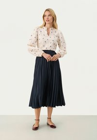 Kremasta bluzica s dolgimi rokavi in mornarsko modrim cvetličnim vzorcem, v kombinaciji z mornarsko modrim plisiranim midi krilom. Model nosi maroon balerinke in trak okoli gležnja.