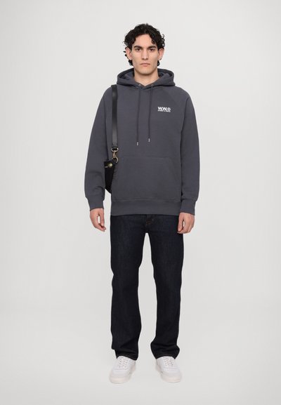 Donkergrijze hoodie met een voorzak, koordjes voor de capuchon en een logo. Gecombineerd met een paar donkere spijkerbroeken en witte sneakers, staand tegen een neutrale achtergrond.