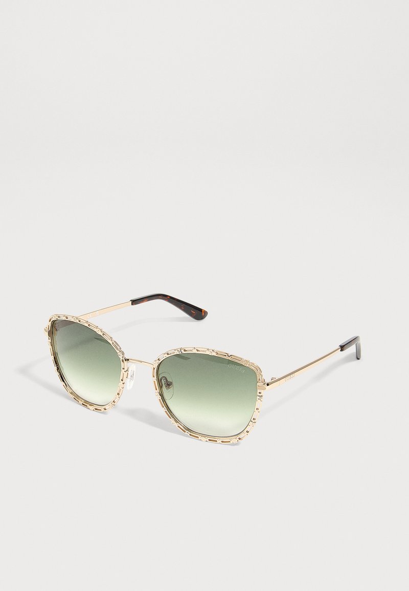 Lunettes de soleil avec une monture métallique dorée aux bords texturés et des verres dégradés verts reposant sur une surface claire.