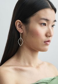 Sølv dråpeøredobber med en scalloped, utsmykket design som fremhever en delikat kjede. Modellen har rett svart hår og et nøytralt uttrykk.