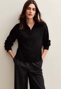 Czarny sweter w dzianinę z kołnierzykiem, luźny krój i ściągacze u rękawów. Połączony z czarno-białymi spodniami w paski o szerokich nogawkach.