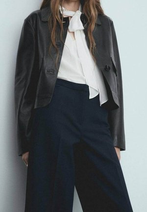 Een zwart leren cropped jack met een knoopsluiting, gecombineerd met een witte blouse met een strikdetail, en marineblauwe nette broek.