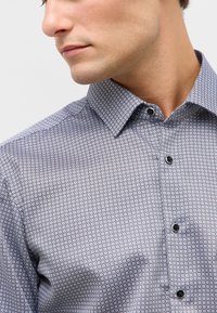 Primo piano di un uomo che indossa una camicia a bottoni con motivo e colletto a punta, rivolto leggermente a sinistra contro uno sfondo semplice.