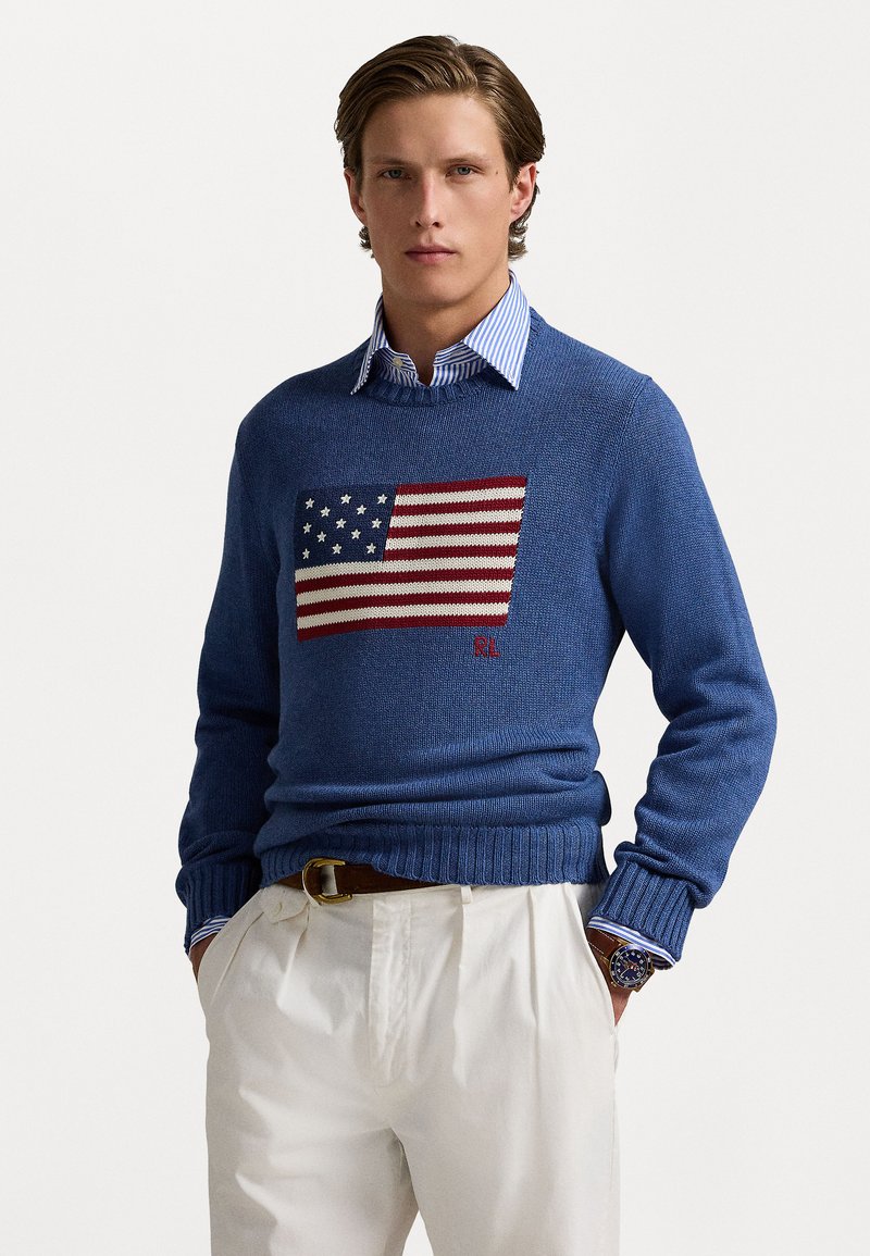 Blue knit sweater featuring a large American flag design on the front, ribbed cuffs and hem, paired with a striped collared shirt and light pants.