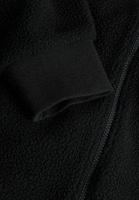 Detail einer schwarzen Fleecejacke, die eine weiche Textur, gerippte Bündchen und einen Reißverschluss aufweist. Der Stoff hat eine plüschige, leicht erhabene Oberfläche.